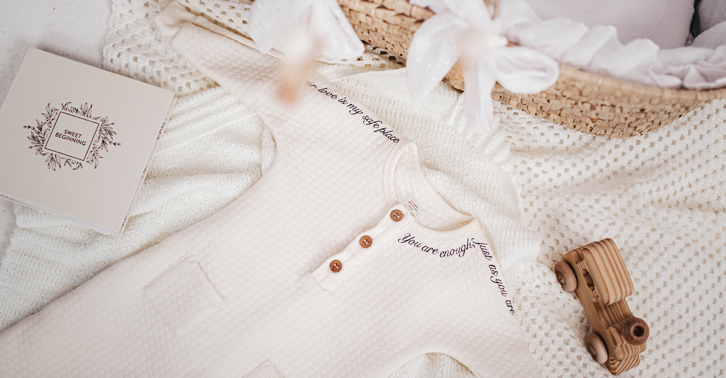 Cream baby romper with affirmation embroidery laid on a knitted blanket beside a wicker basket and wooden toy car