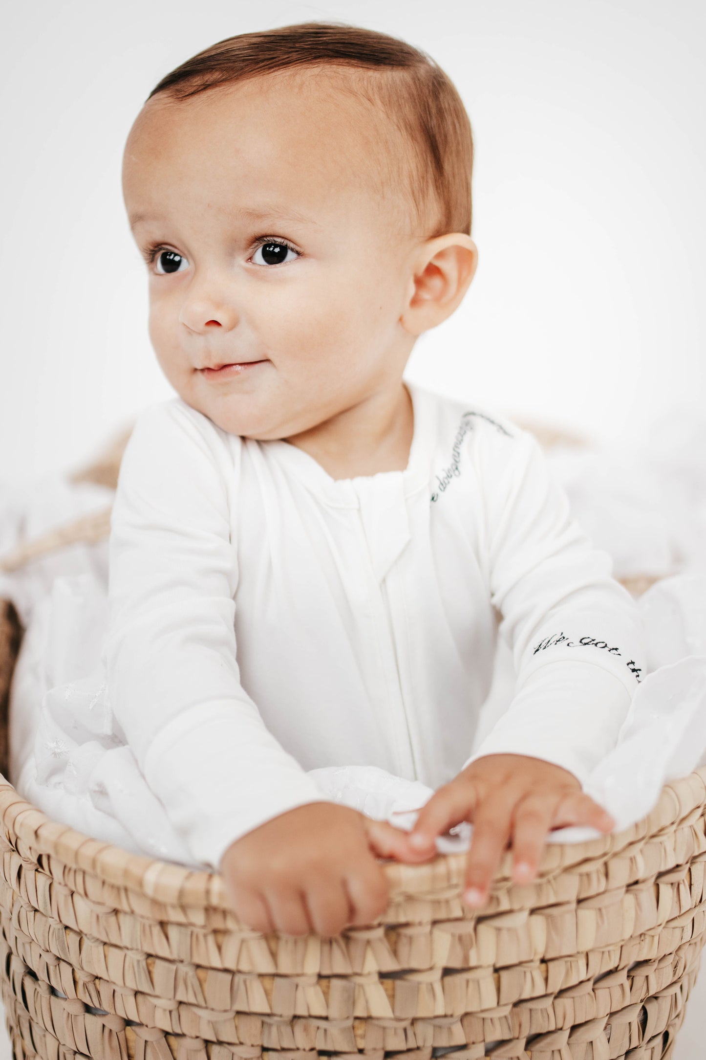 Baby in a white outfit sitting in a woven basket on a white background