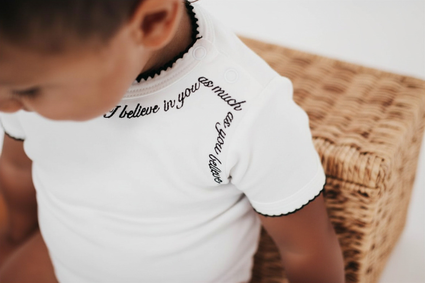 Child wearing a white shirt with text, sitting on a wicker chair.